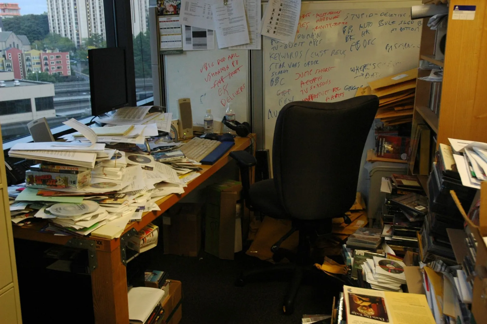 A cluttered office desk buried in papers, discs, and scribbled whiteboard notes