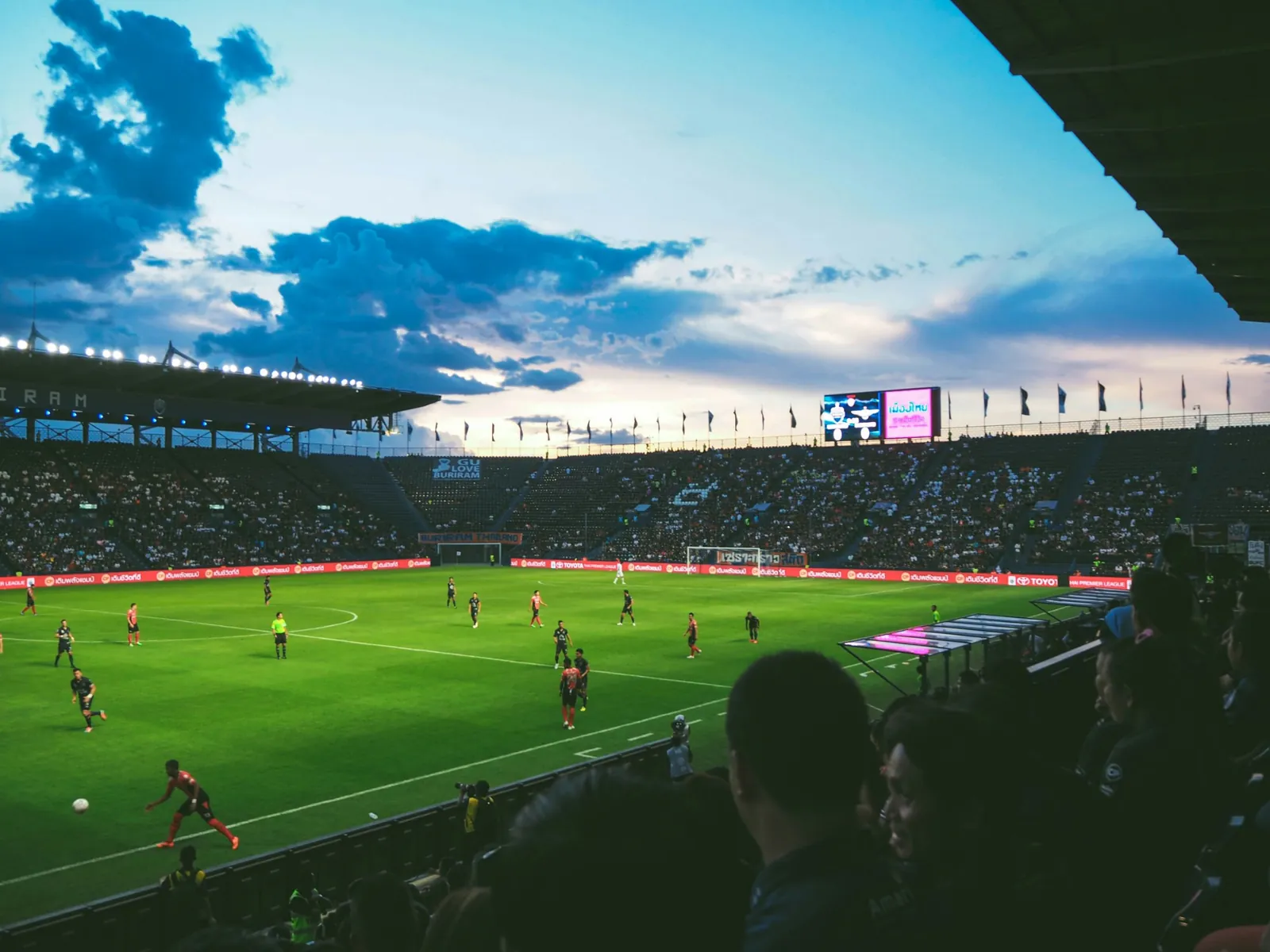 A packed football stadium at dusk with players on the pitch under floodlights